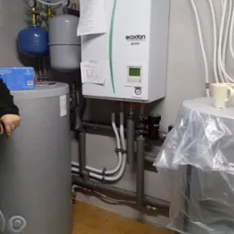 Biały piec gazowy Ecodan Mitsubishi Electric z szarym zbiornikiem, rurami i osprzętem w kotłowni, widoczne zabezpieczenia rur.