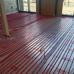 Usługi Hydrauliczne Mateusz Jarzembowski - Czerwone rury ogrzewania podłogowego ułożone na izolacji z siatką w nowym budynku z dużymi oknami. Widoczne elementy konstrukcyjne budynku.