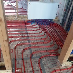 Usługi Hydrauliczne Mateusz Jarzembowski - Czerwone rury ogrzewania podłogowego, zamocowane na folii z siatką, widoczne drewniane elementy konstrukcyjne i biała skrzynka rozdzielcza. Instalacja w trakcie montażu.