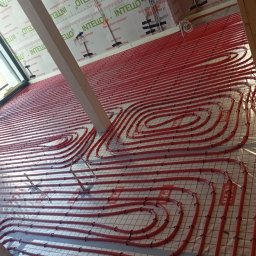 Usługi Hydrauliczne Mateusz Jarzembowski - Czerwone rury ogrzewania podłogowego ułożone na siatce, widoczne elementy konstrukcyjne budynku i izolacja. Instalacja przed wylaniem posadzki.