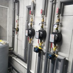 Usługi Hydrauliczne Mateusz Jarzembowski - Szare, zaizolowane rury instalacji grzewczej z zaworami i manometrami, zamontowane na kafelkowanej ścianie w kotłowni. Widoczny fragment zbiornika.