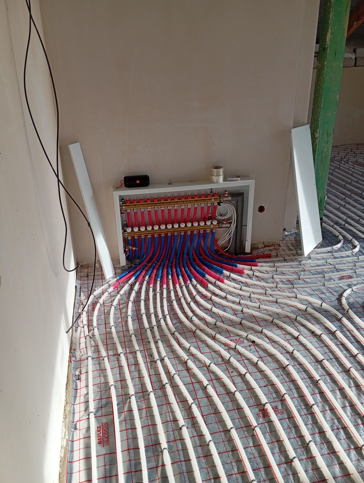 Rozdzielacz z rurami ogrzewania podłogowego, ułożonymi na izolacji z folią w kratkę. Rury czerwone i niebieskie. Widoczne kable i elementy konstrukcyjne.