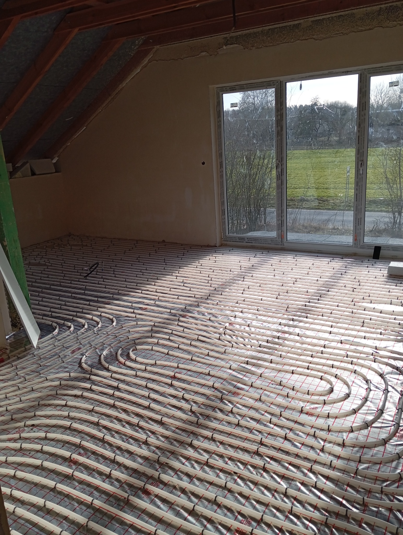 Białe rury ogrzewania podłogowego ułożone na izolacji w jasnym pokoju z dużym oknem, widok na zielone pole. Instalacja w trakcie realizacji, widoczne elementy konstrukcyjne.