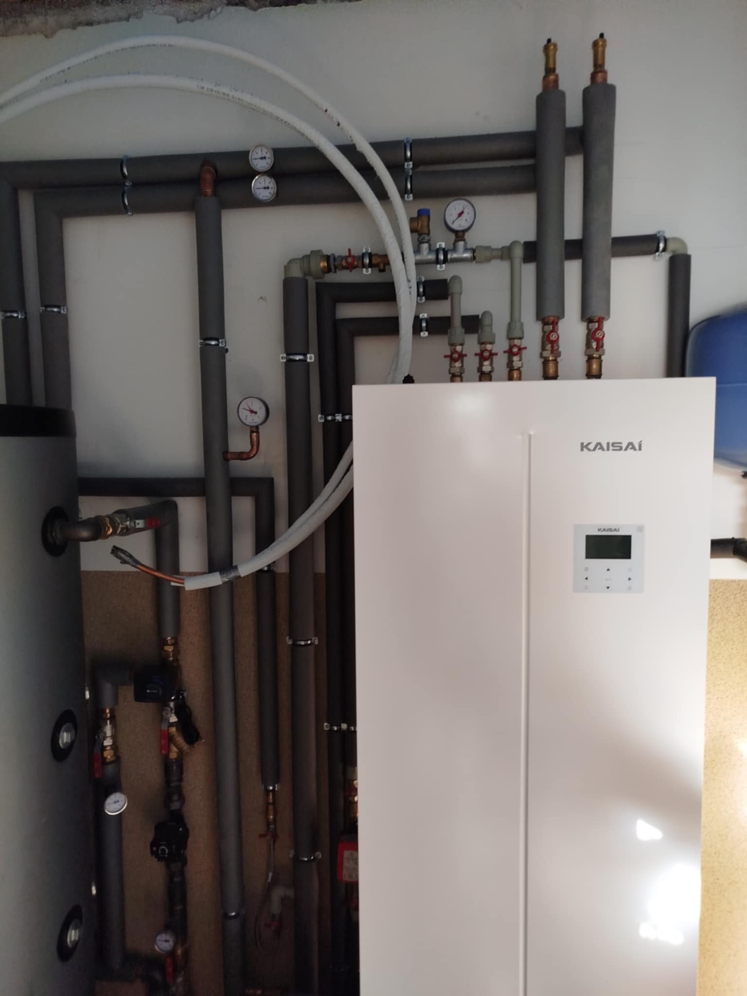 Biała pompa ciepła Kaisai z podłączonymi rurami, zaworami i manometrami. Widoczne izolowane rury grzewcze i zasobnik wody. Nowoczesna instalacja grzewcza w kotłowni.