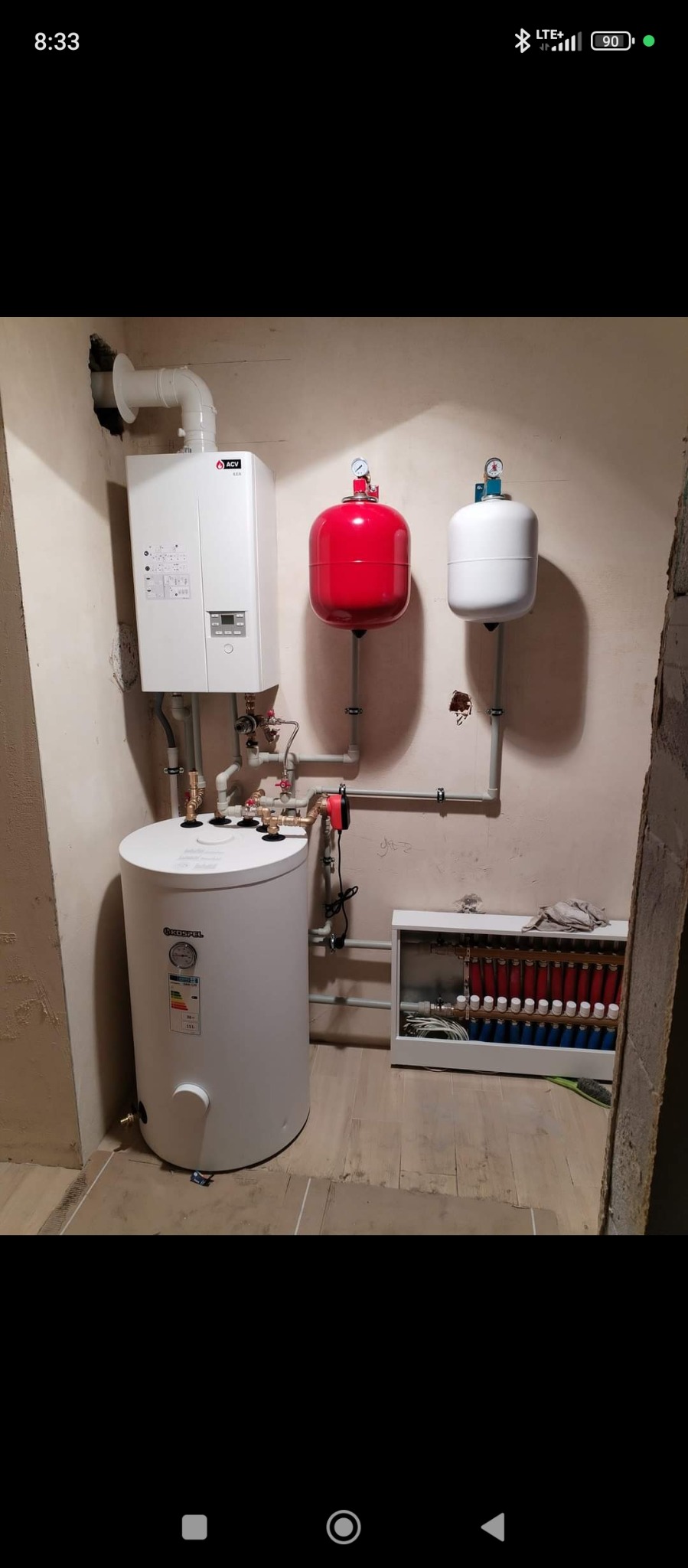 Kotłownia z białym bojlerem Kospel, piecem ACV, czerwonym i białym zbiornikiem wyrównawczym. Rozdzielacz ogrzewania podłogowego w szafce.