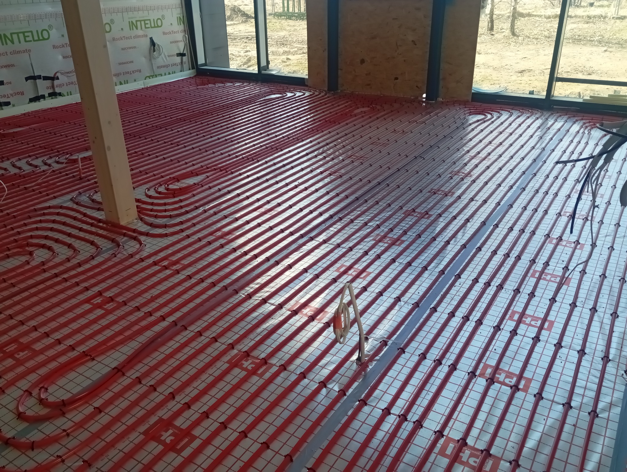 Czerwone rury ogrzewania podłogowego ułożone na izolacji z siatką w nowym budynku z dużymi oknami. Widoczne elementy konstrukcyjne budynku.