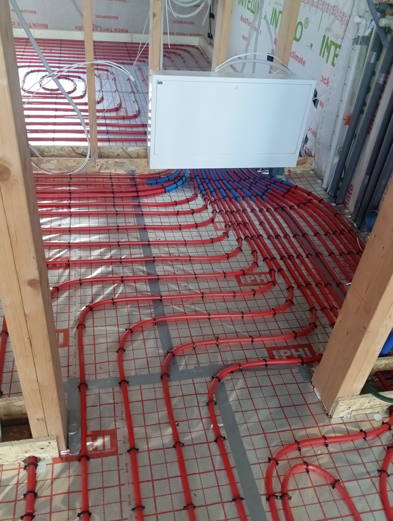 Czerwone rury ogrzewania podłogowego, zamocowane na folii z siatką, widoczne drewniane elementy konstrukcyjne i biała skrzynka rozdzielcza. Instalacja w trakcie montażu.