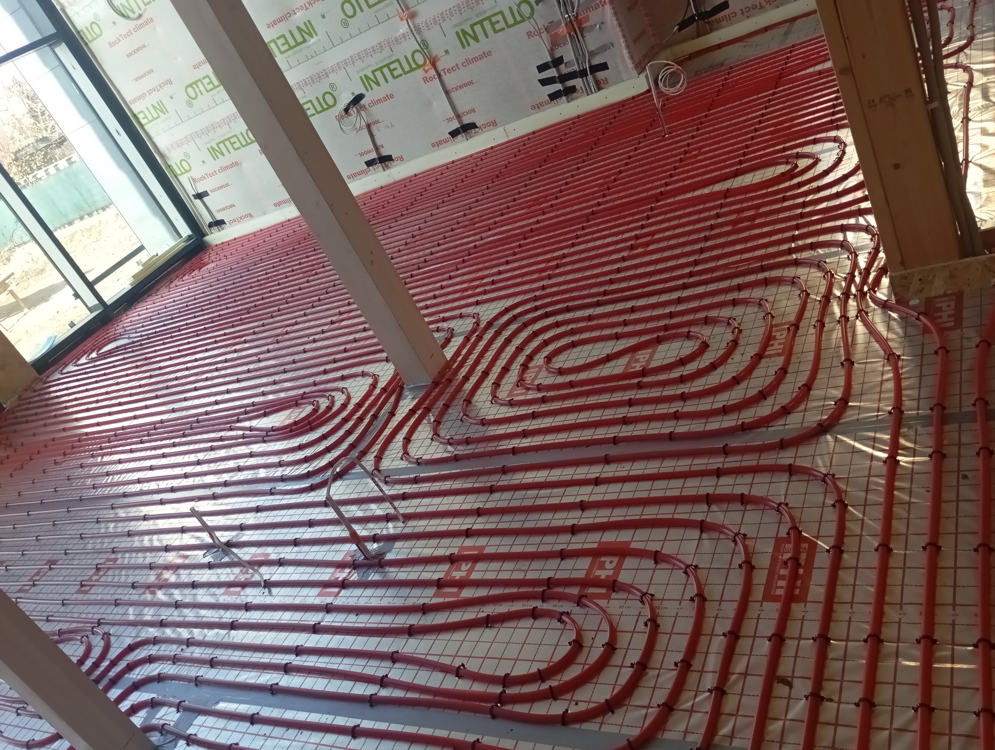 Czerwone rury ogrzewania podłogowego ułożone na siatce, widoczne elementy konstrukcyjne budynku i izolacja. Instalacja przed wylaniem posadzki.