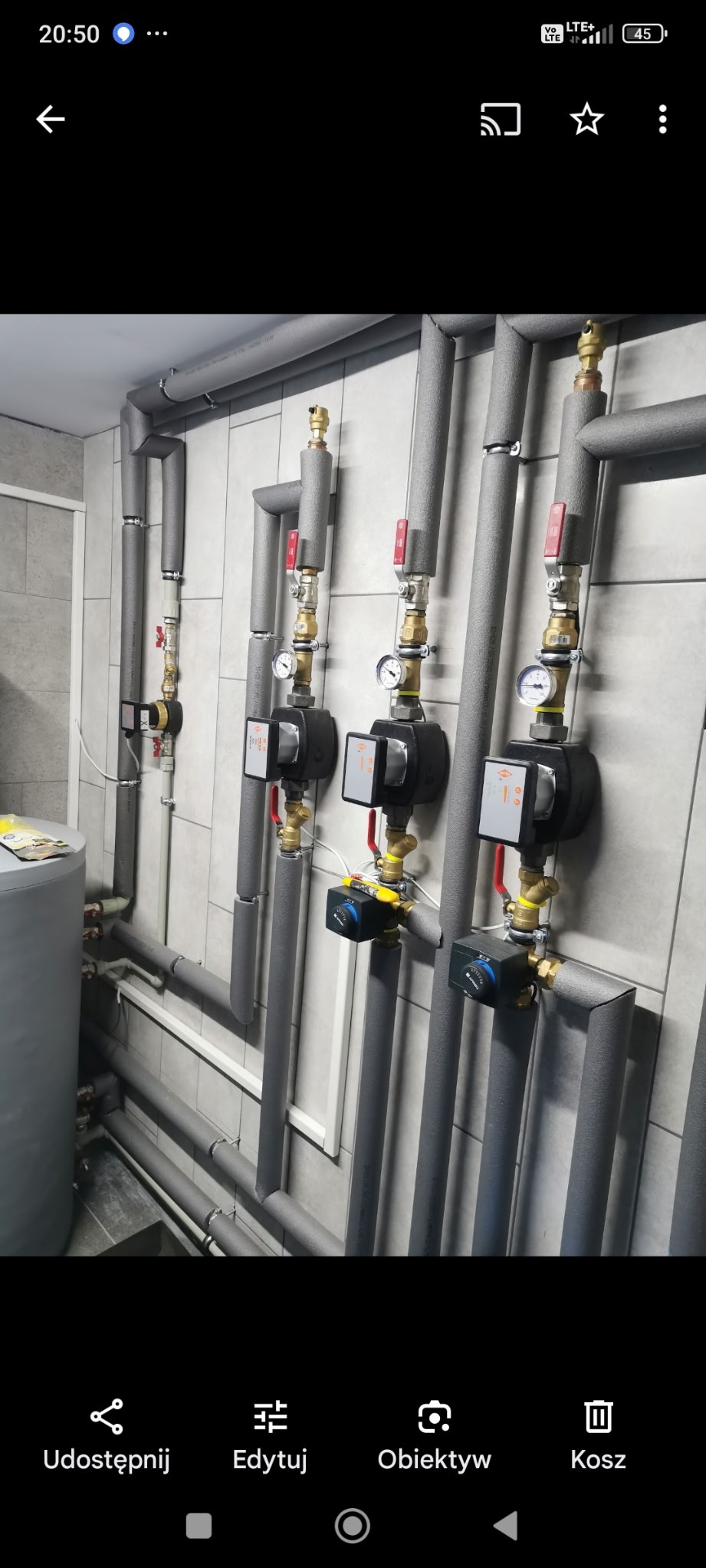 Szare, zaizolowane rury instalacji grzewczej z zaworami i manometrami, zamontowane na kafelkowanej ścianie w kotłowni. Widoczny fragment zbiornika.