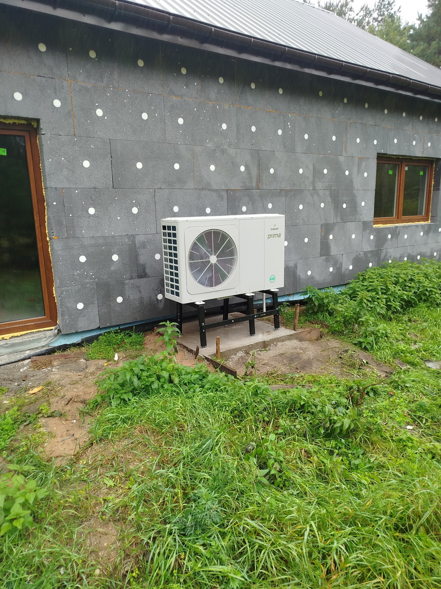 Biała pompa ciepła zamontowana na zewnątrz budynku z szarą elewacją, widoczne okna z brązowymi ramami i zielona trawa. Instalacja na betonowej podstawie.