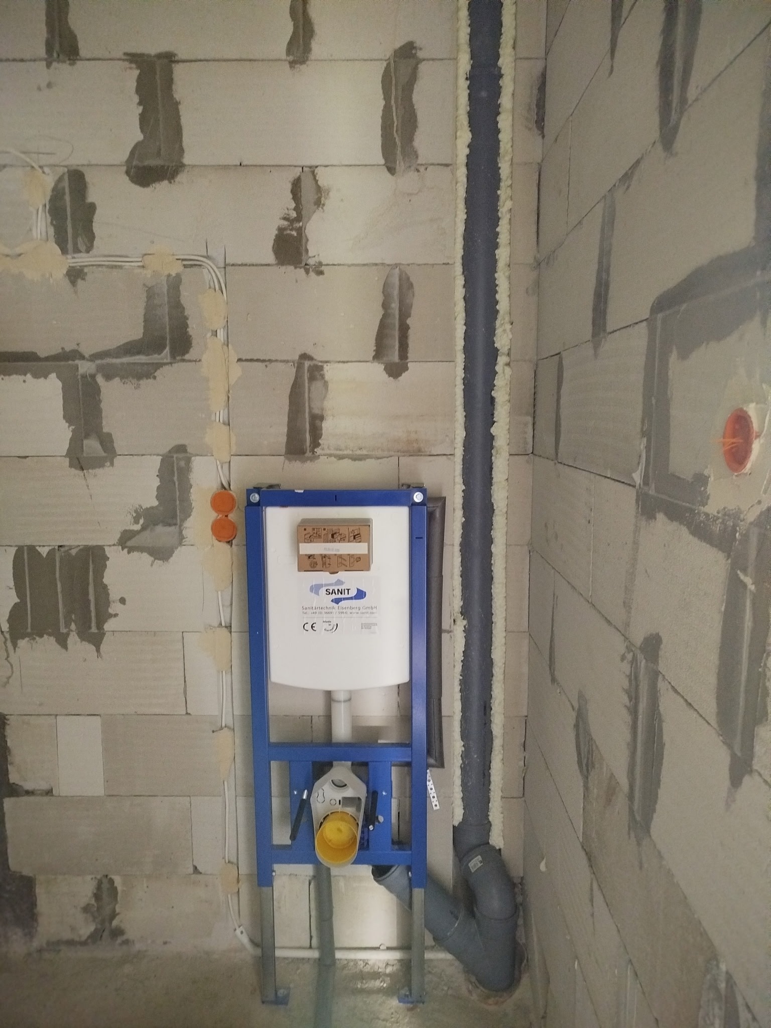 Montaż stelaża podtynkowego WC marki Sanit na surowej ścianie z betonu komórkowego. Widoczne rury kanalizacyjne i instalacja elektryczna w ścianie.