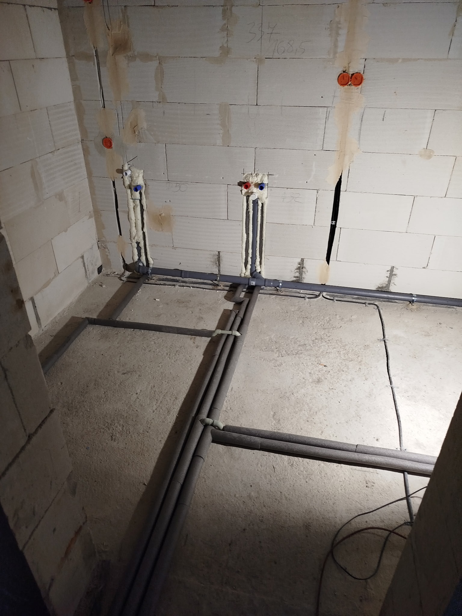 Surowy stan: instalacja wodno-kanalizacyjna z rurami w izolacji na podłodze i wyprowadzeniami ze ściany z bloków. Widoczne puszki elektryczne.