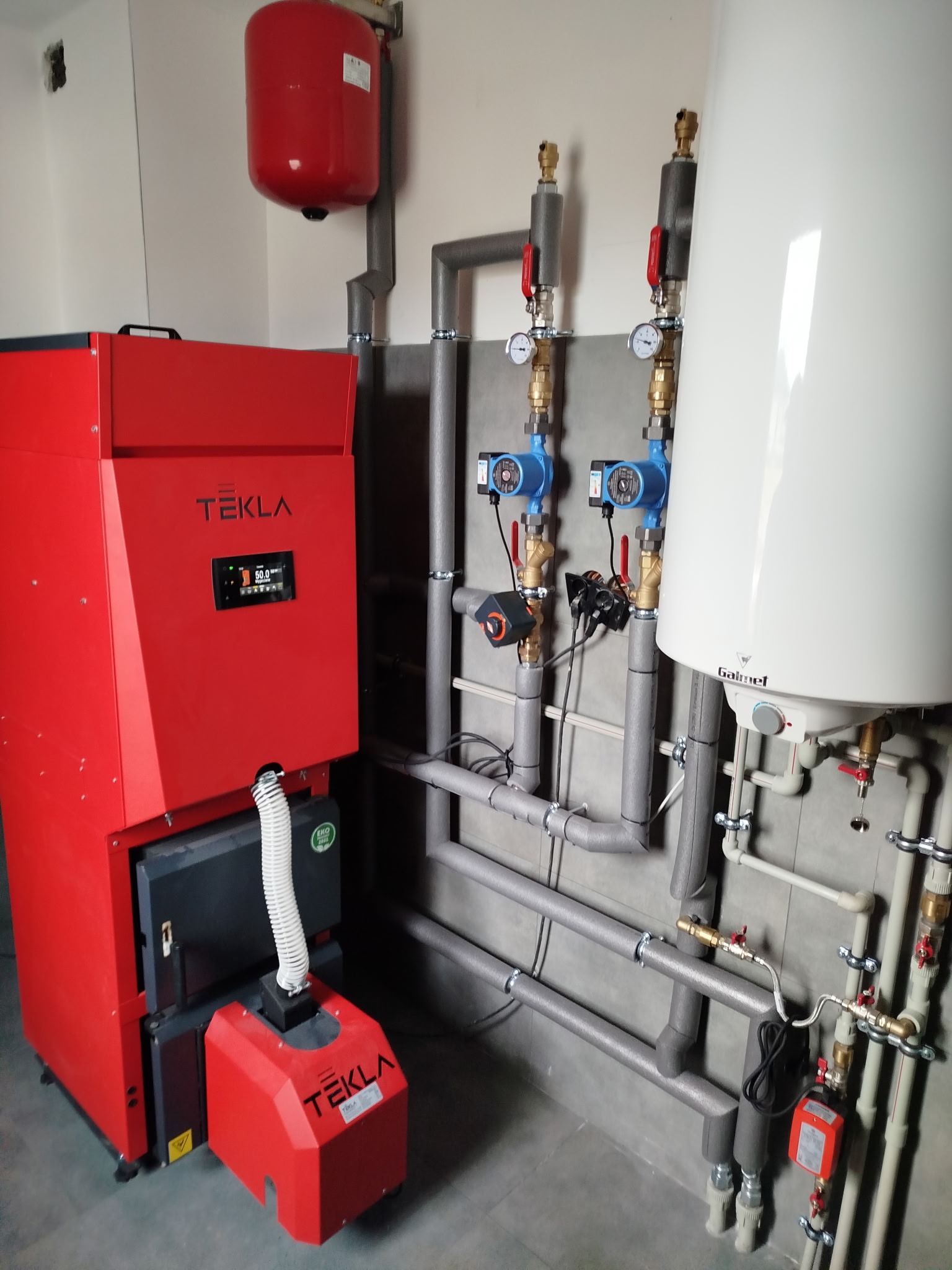 Czerwony piec Tekla z podajnikiem, biały bojler Galmet i szare rury instalacji grzewczej na tle szarej ściany. Widoczne pompy obiegowe i zawory.