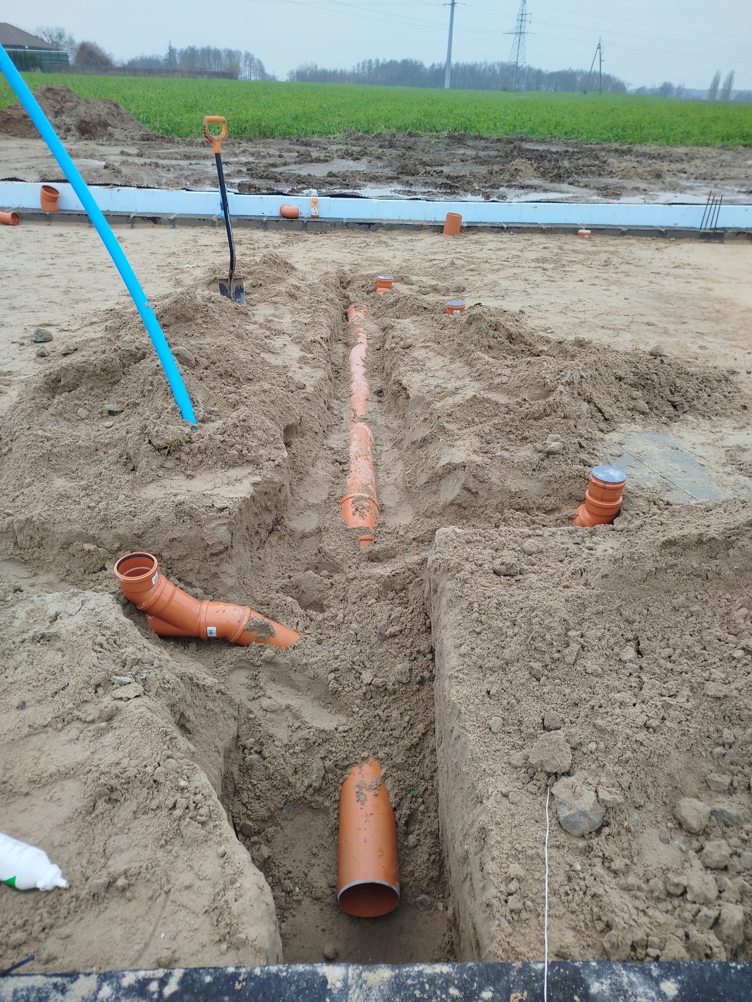 Wykop z ułożonymi rurami kanalizacyjnymi z tworzywa sztucznego w piasku, widoczna łopata i niebieska rura. W tle zielone pole i słupy energetyczne.