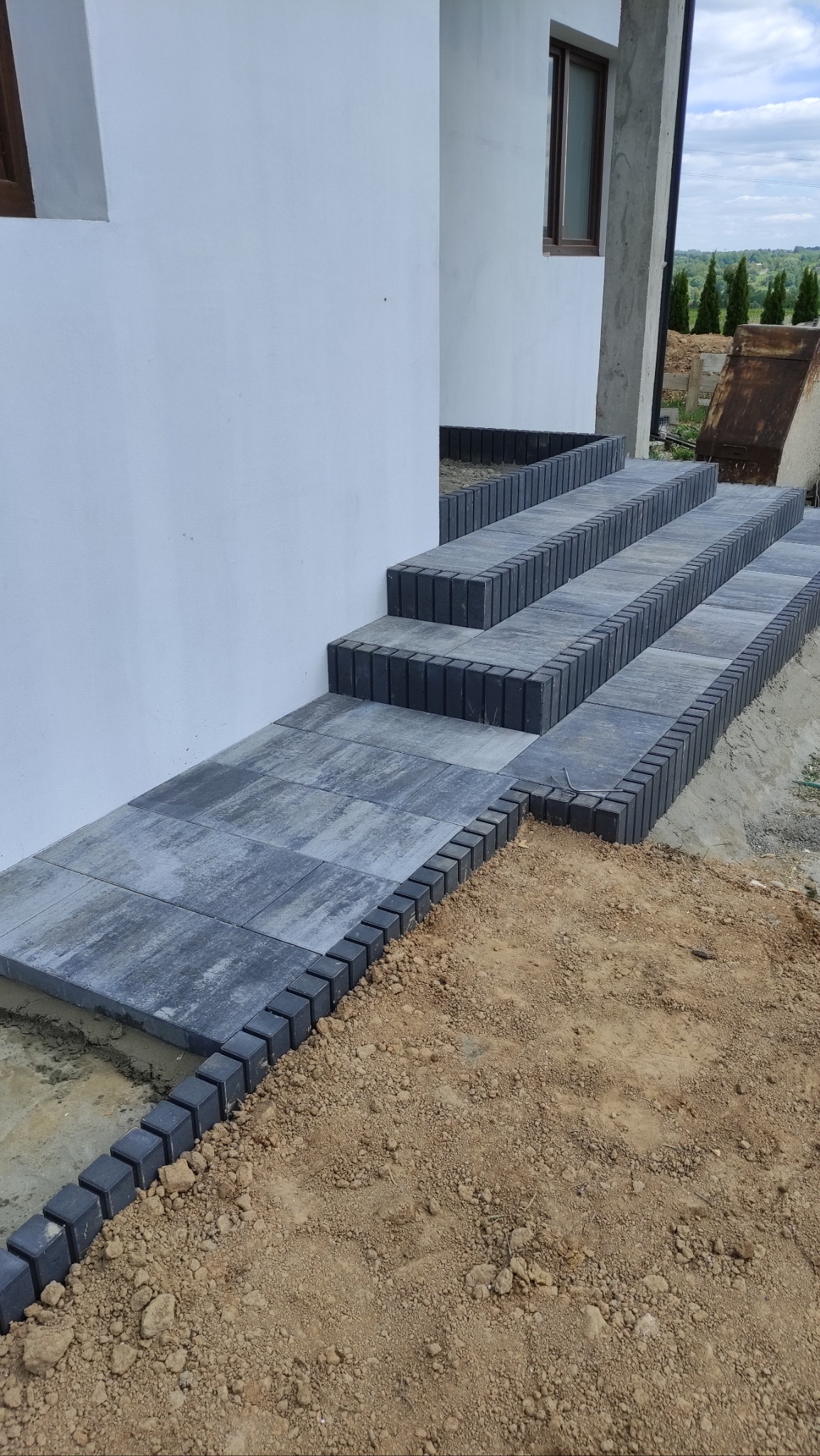 Ułożone schody i chodnik z szarej kostki brukowej obrzeżonej czarnym krawężnikiem, prowadzące do budynku z białą elewacją. Widoczne ślady prac ziemnych.