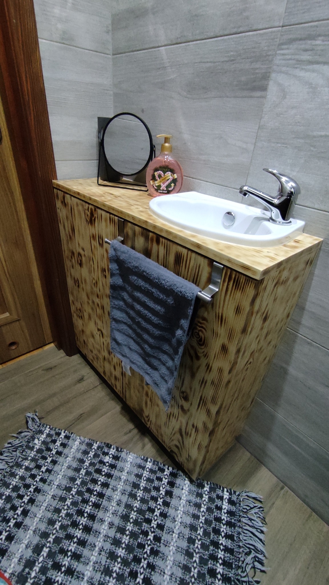Nowoczesna łazienka z drewnianą szafką pod umywalką, lustrem i ręcznikiem. Szafka z widocznym usłojeniem drewna. Szare płytki na ścianie i dywanik na podłodze.