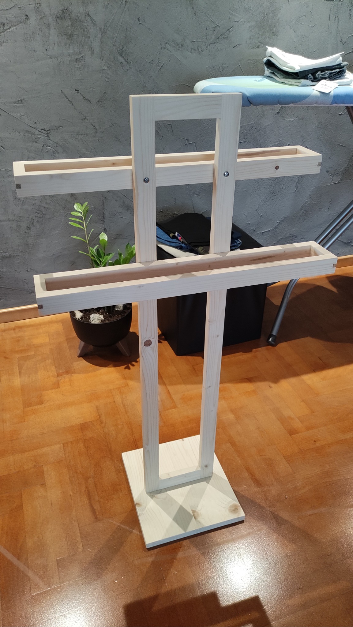 Surowy, drewniany regał z półkami na tle szarej ściany i parkietu. Widoczny element deski do prasowania i kwiat doniczkowy. Mebel w stylu skandynawskim.