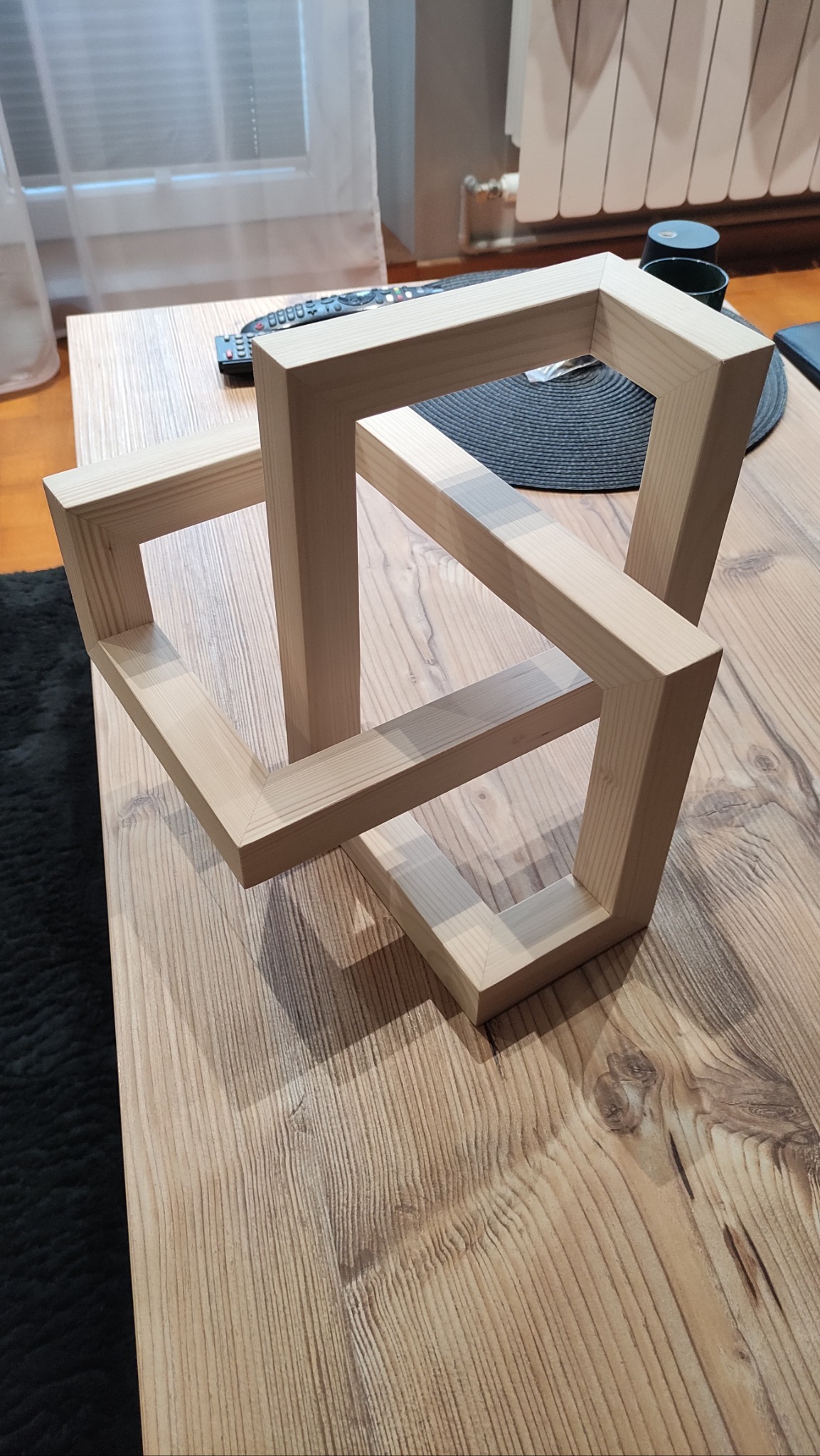 Drewniana, przestrzenna konstrukcja z jasnego drewna, ustawiona na stole o wyraźnym rysunku słojów. W tle widoczne okno z zasłoną i fragment grzejnika.