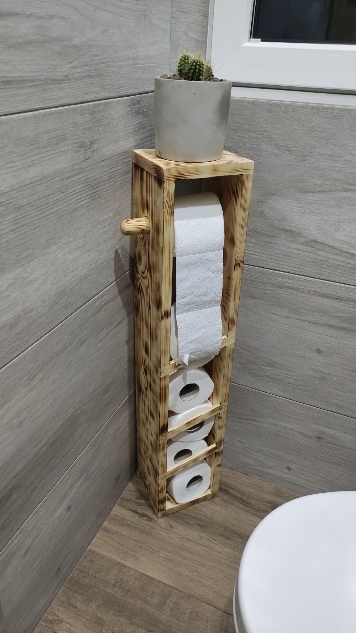 Drewniany regał na papier toaletowy, z kaktusem na górze, stoi w łazience z szarymi płytkami imitującymi drewno, obok widoczny fragment toalety.