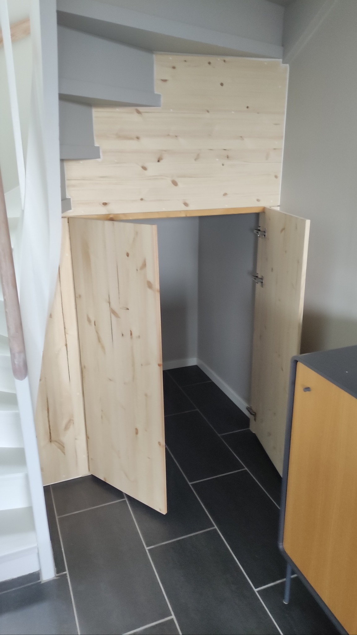 Drewniana zabudowa pod schodami z otwartymi drzwiczkami, widoczna czarna podłoga z płytek i fragment schodów. Naturalne drewno, proste wykończenie.