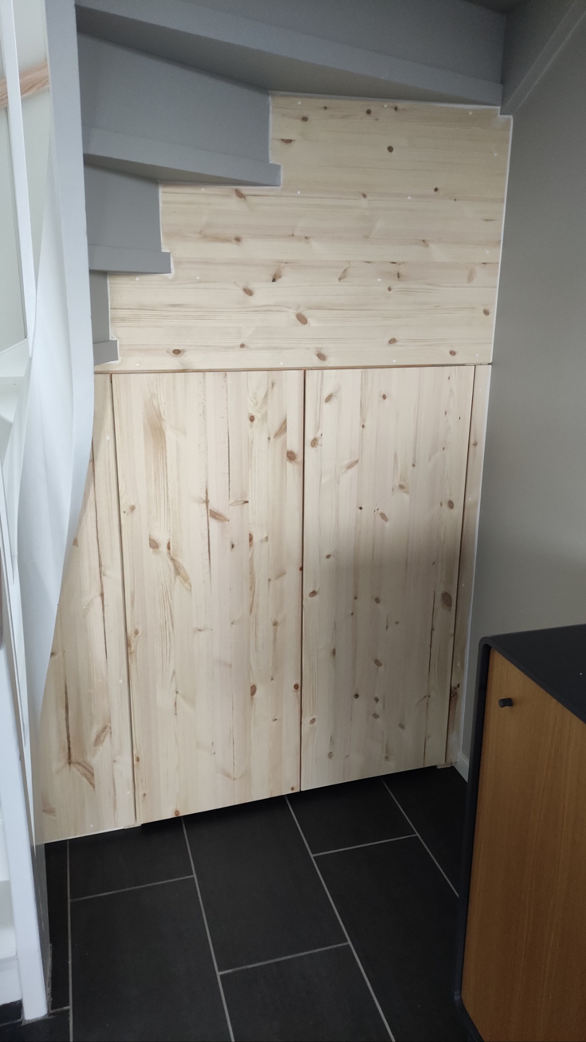 Drewniana zabudowa pod schodami z czterema drzwiczkami, wykończona boazerią. Ciemna, prostokątna podłoga i fragment schodów z boku.