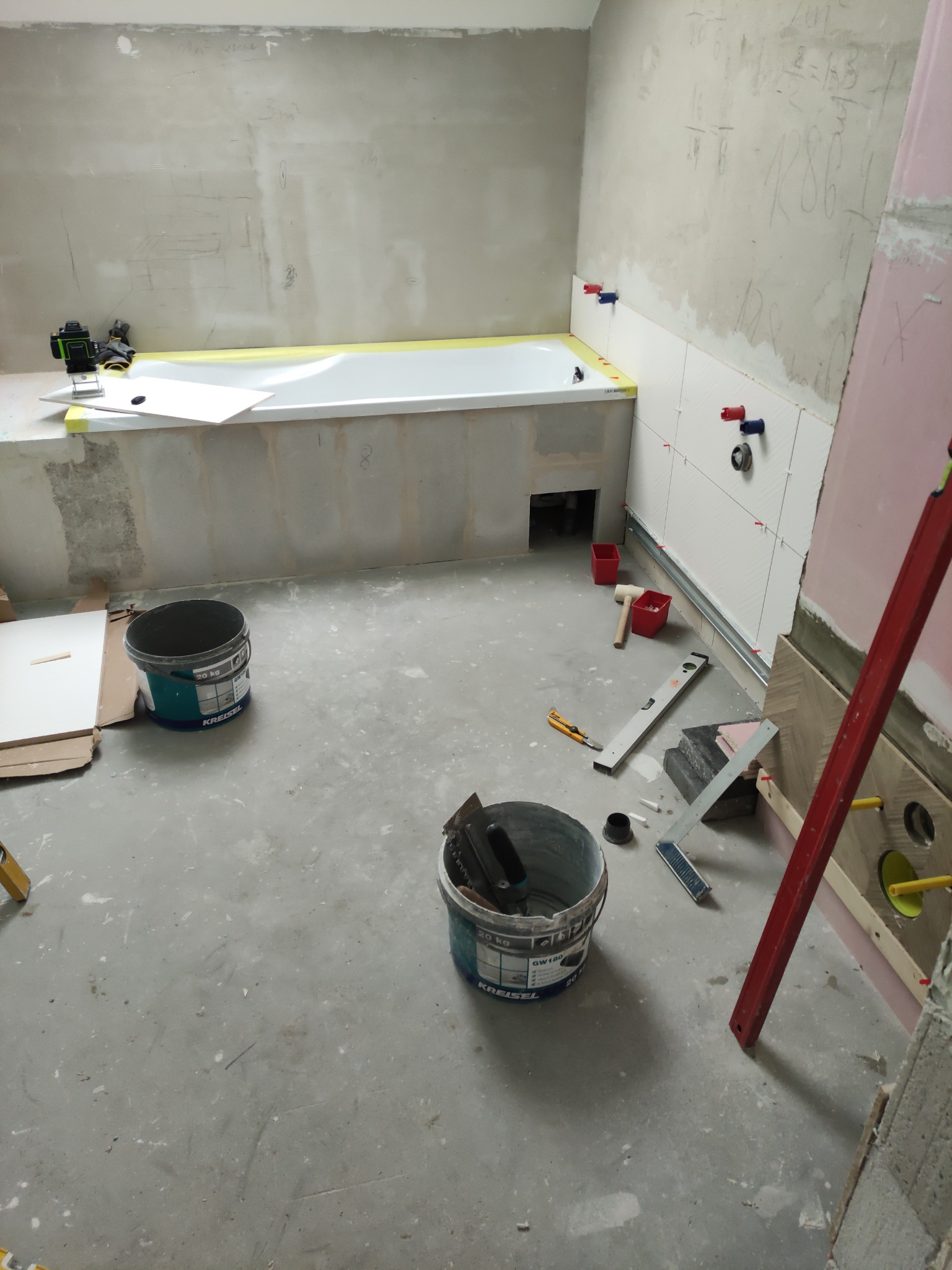 Remont łazienki: wanna obudowana, częściowo wyłożona płytkami. Widoczne narzędzia, poziomica, wiaderka z zaprawą. Fragment instalacji wodnej.
