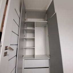 Marcin Kamiński - Otwarta, biało-szara szafa z półkami i szufladami, widok z dołu, drzwi z dekoracyjnymi frezowaniami, srebrne uchwyty, wnętrze szafy podzielone na segmenty.