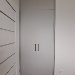 Marcin Kamiński - Zamknięta, szara szafa wnękowa z długimi, czarnymi uchwytami na tle białej ściany. Po lewej stronie widoczny fragment białej szafy z poziomymi frezami.