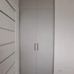 Zamknięta, szara szafa wnękowa z długimi, czarnymi uchwytami na tle białej ściany. Po lewej stronie widoczny fragment białej szafy z poziomymi frezami.