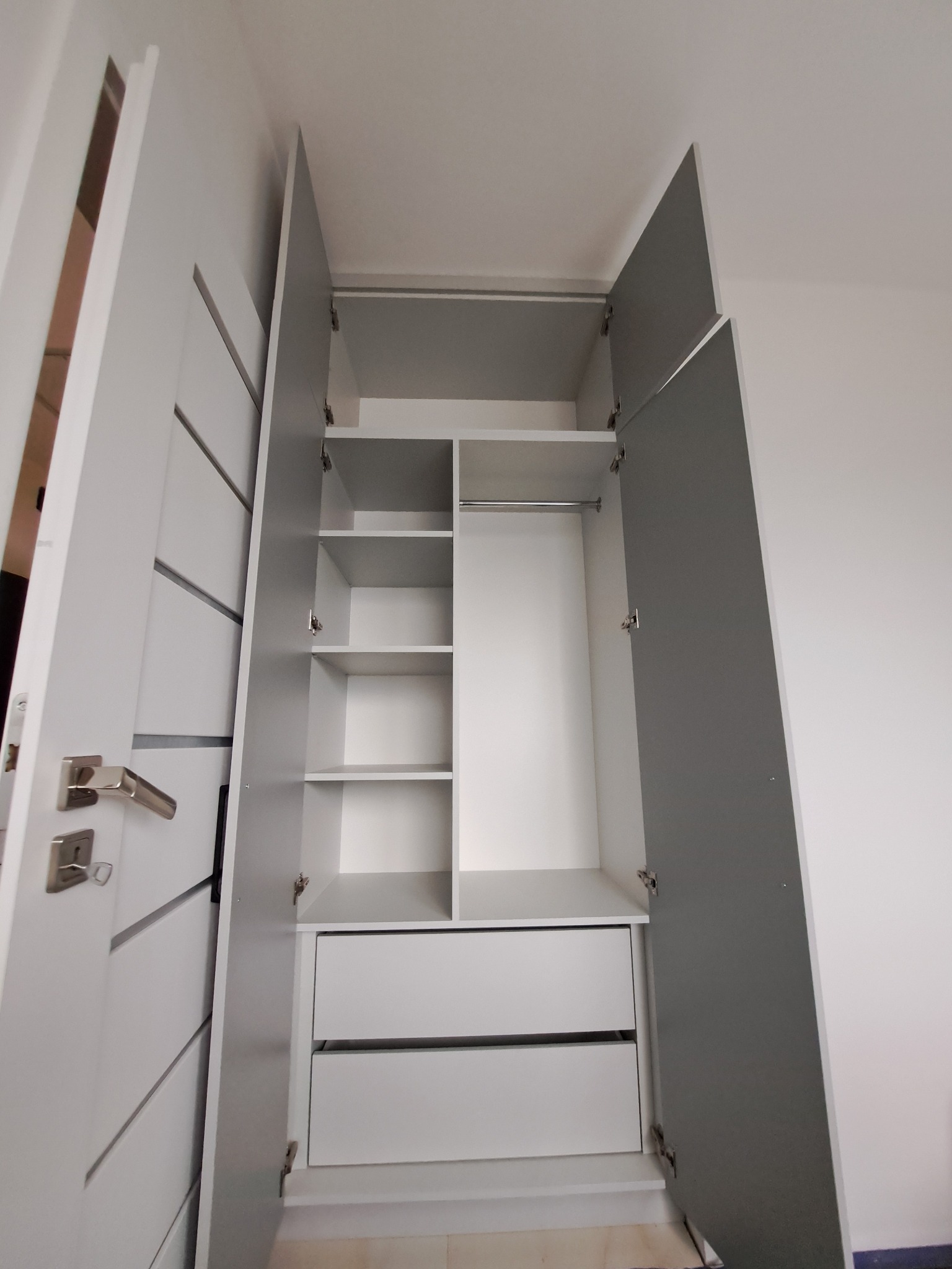 Otwarta, biało-szara szafa z półkami i szufladami, widok z dołu, drzwi z dekoracyjnymi frezowaniami, srebrne uchwyty, wnętrze szafy podzielone na segmenty.