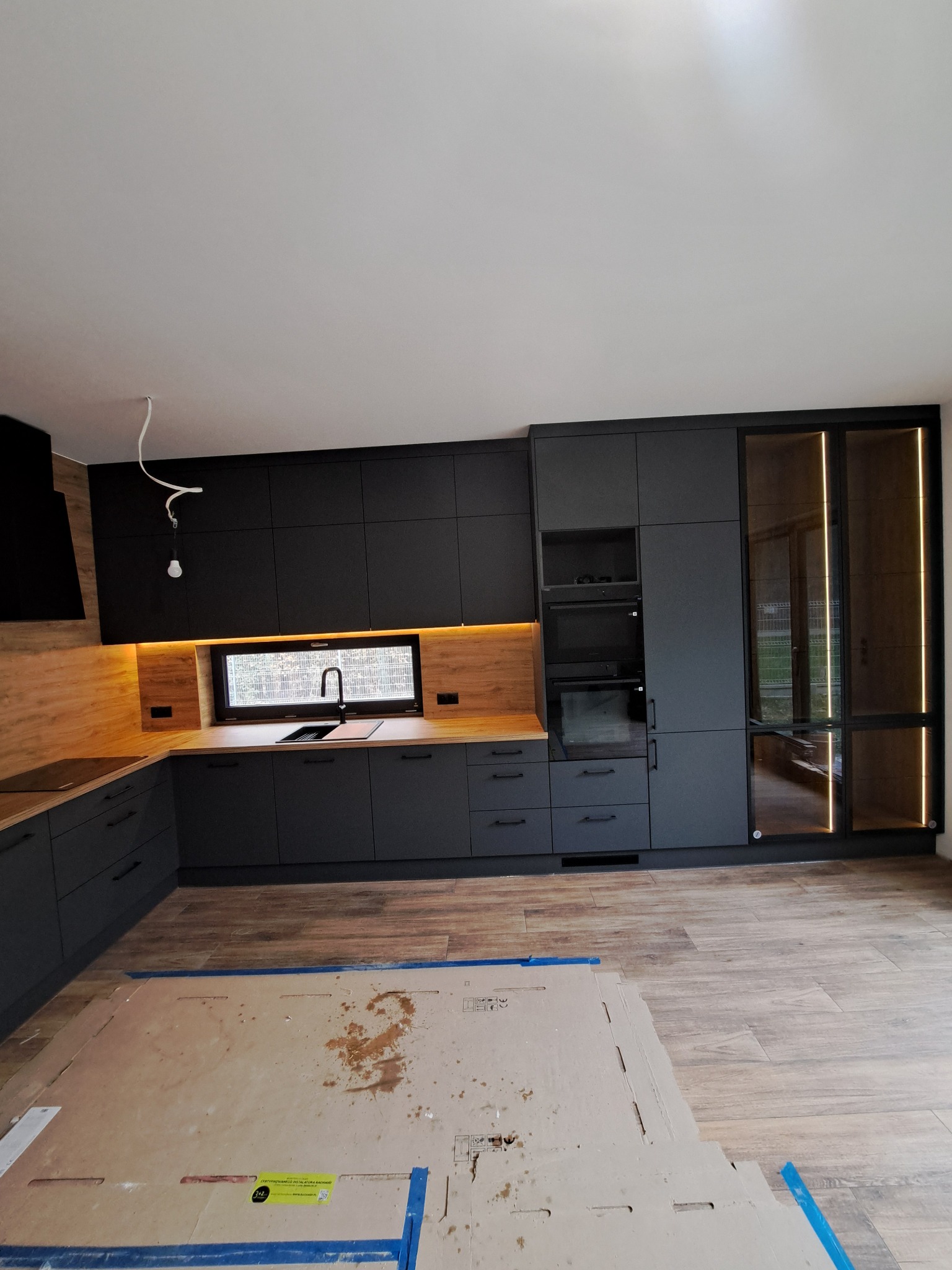 Nowoczesna kuchnia z czarnymi matowymi frontami, drewnianym blatem i oświetleniem LED pod szafkami. Widoczny fragment podłogi zabezpieczony kartonem.