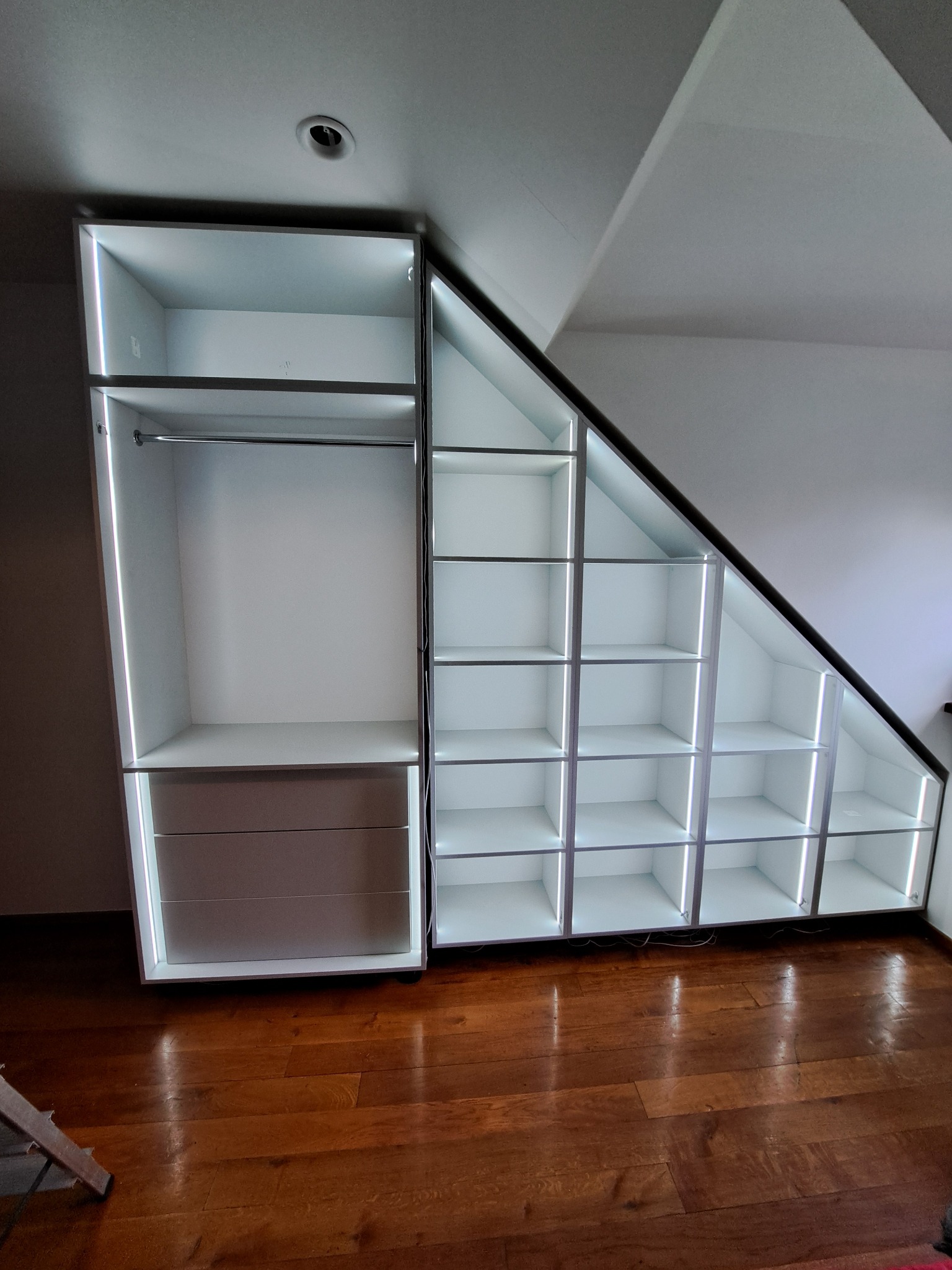 Biała szafa wnękowa z oświetleniem LED, dopasowana do skosu dachu. Widoczne półki i szuflady, drewniana podłoga. Nowoczesny design.