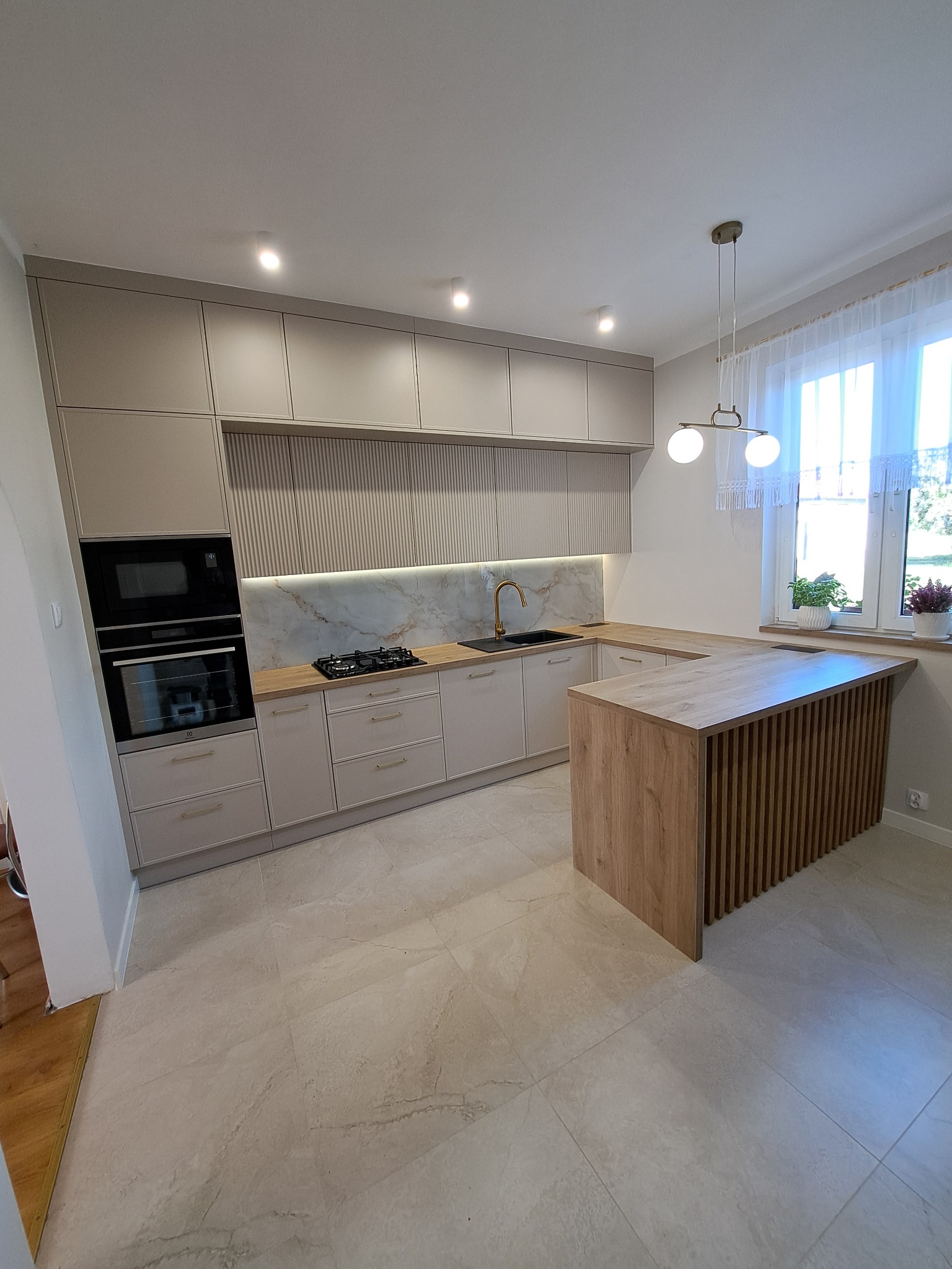 Jasna kuchnia z wyspą i ryflowanymi frontami szafek. Złota armatura i marmurowy backsplash dodają elegancji. Widoczne okno z roślinami i nowoczesna lampa.