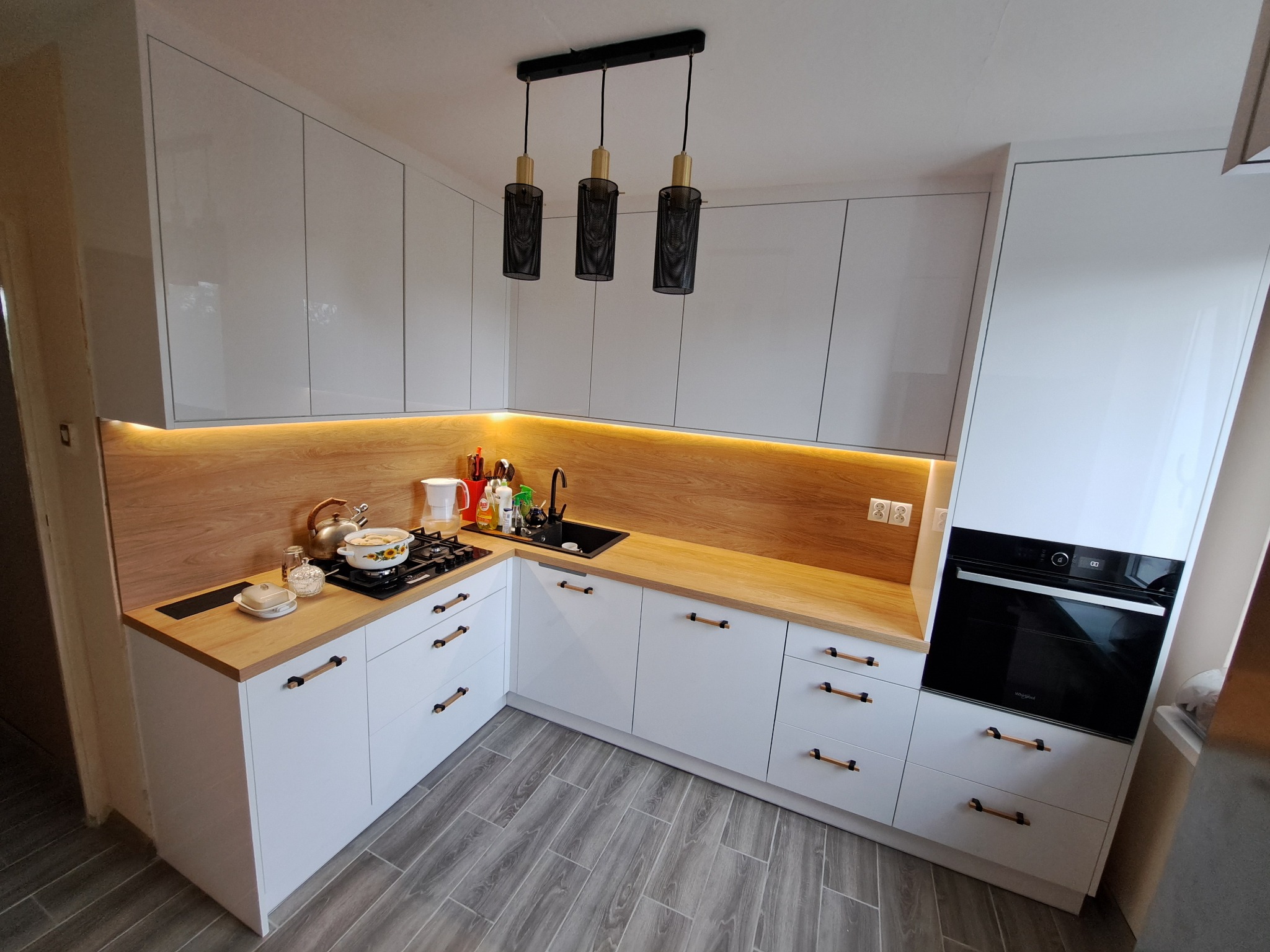 Biała kuchnia na wymiar z drewnianym blatem i czarnymi lampami wiszącymi. Widoczne AGD i akcesoria kuchenne. Podświetlenie blatu dodaje ciepła.