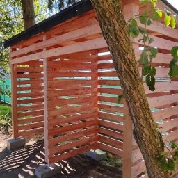 SŁOWIK WOOD - Drewniana konstrukcja z poziomych desek, prawdopodobnie wiata na drewno, z czarnym dachem, wsparta na betonowych bloczkach, obok pnia drzewa, na tle zieleni.