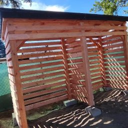 SŁOWIK WOOD - Nowa, drewniana konstrukcja z listew, prawdopodobnie wiata, w naturalnym otoczeniu z drzewami i błękitnym niebem w tle, widziana z boku.