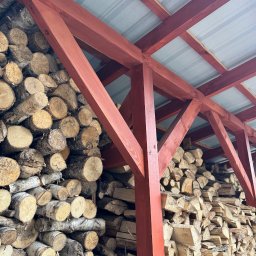 SŁOWIK WOOD - Czerwona, drewniana konstrukcja z zadaszeniem, pod którą składowane jest drewno opałowe. Widoczne pocięte kawałki drewna ułożone w stos.