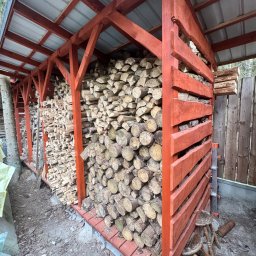 SŁOWIK WOOD - Duża drewutnia z czerwonymi, drewnianymi elementami konstrukcyjnymi i metalowym dachem, wypełniona pociętym drewnem, z dwoma stołkami obok.