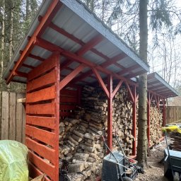 SŁOWIK WOOD - Drewniana wiata na drewno z szarą blachą falistą, wypełniona pociętymi klockami drewna. Obok taczka i narzędzia. W tle las i ogrodzenie.