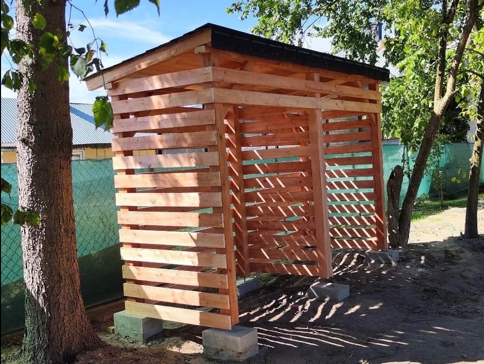 Drewniana drewutnia z poziomych desek, z czarnym dachem, umieszczona na betonowych bloczkach, widoczna w ogrodzie w słoneczny dzień.