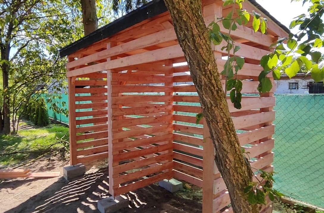 Drewniana konstrukcja z poziomych desek, prawdopodobnie wiata na drewno, z czarnym dachem, wsparta na betonowych bloczkach, obok pnia drzewa, na tle zieleni.