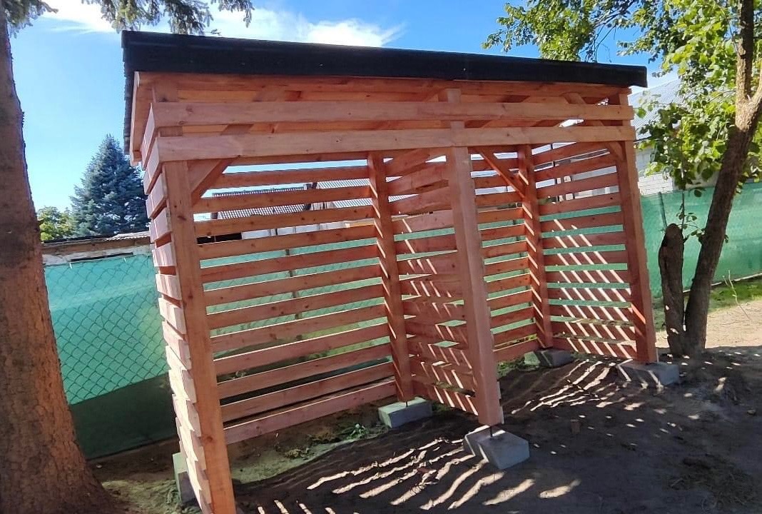 Nowa, drewniana konstrukcja z listew, prawdopodobnie wiata, w naturalnym otoczeniu z drzewami i błękitnym niebem w tle, widziana z boku.