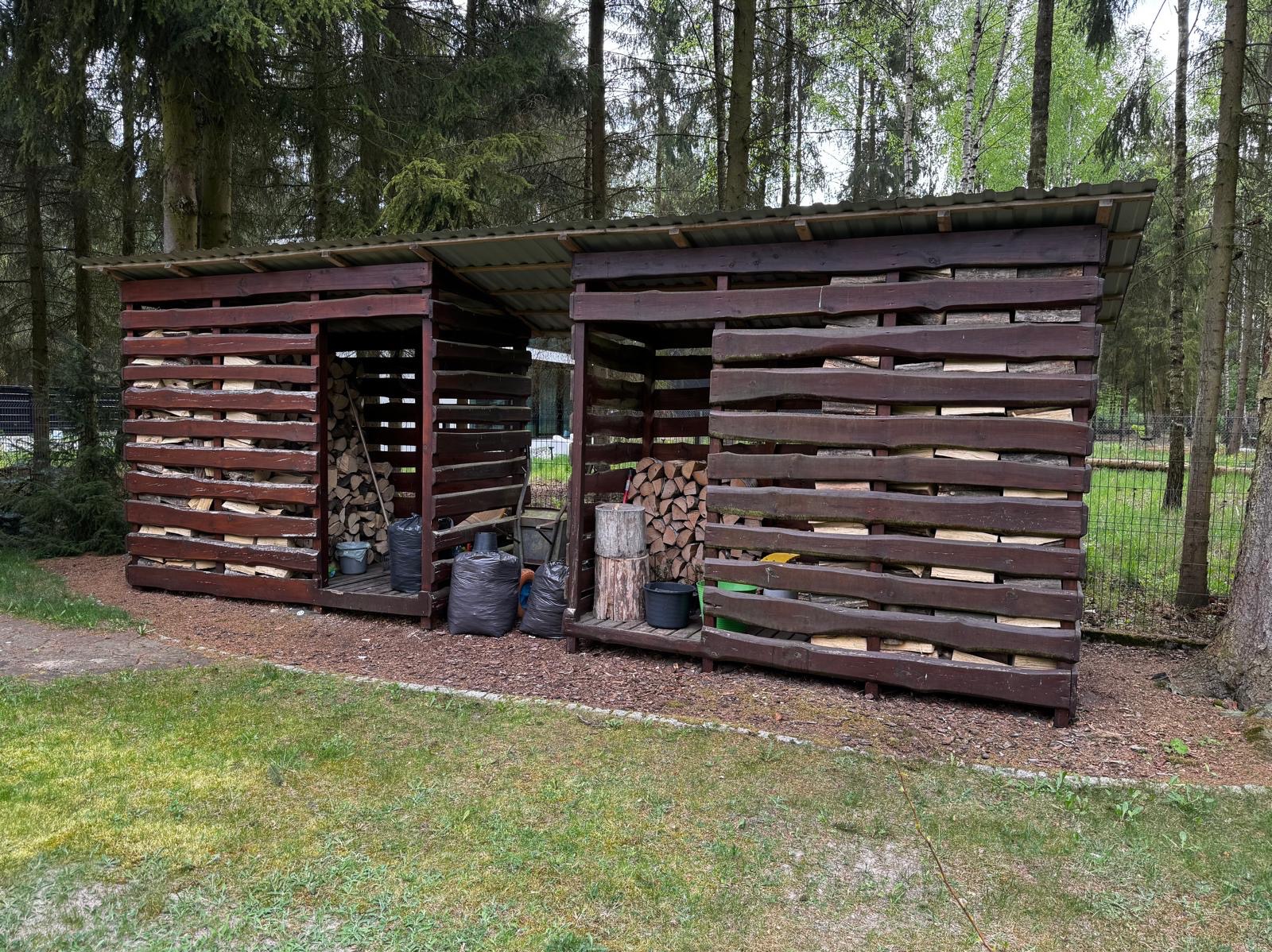 Drewniana konstrukcja z dwoma zadaszonymi przegrodami, wypełniona drewnem opałowym, otoczona drzewami i zielenią. Widoczne worki i narzędzia.