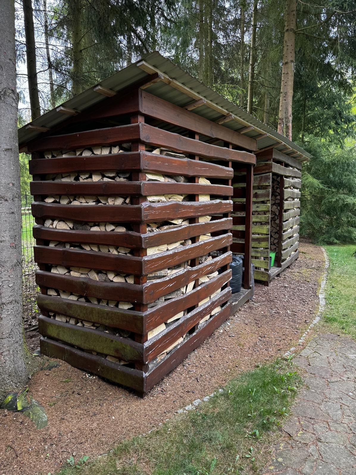 Drewniana konstrukcja do przechowywania drewna opałowego z metalowym dachem, usytuowana w leśnym otoczeniu, widoczna z boku, z elementami mchu na dolnych deskach.