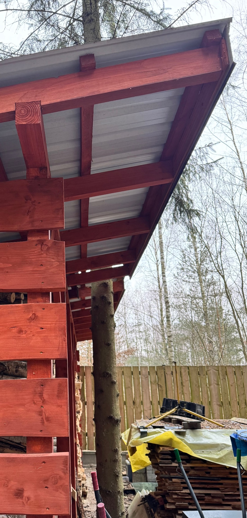 Czerwona drewniana konstrukcja z metalowym dachem, wsparta na pniu drzewa, widok z dołu, w tle ogrodzenie i drzewa bez liści. Konstrukcja z wypełnieniem drewnem.