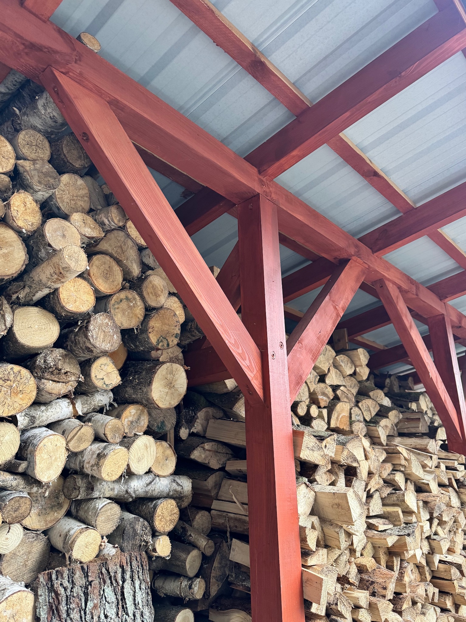 Czerwona, drewniana konstrukcja z zadaszeniem, pod którą składowane jest drewno opałowe. Widoczne pocięte kawałki drewna ułożone w stos.