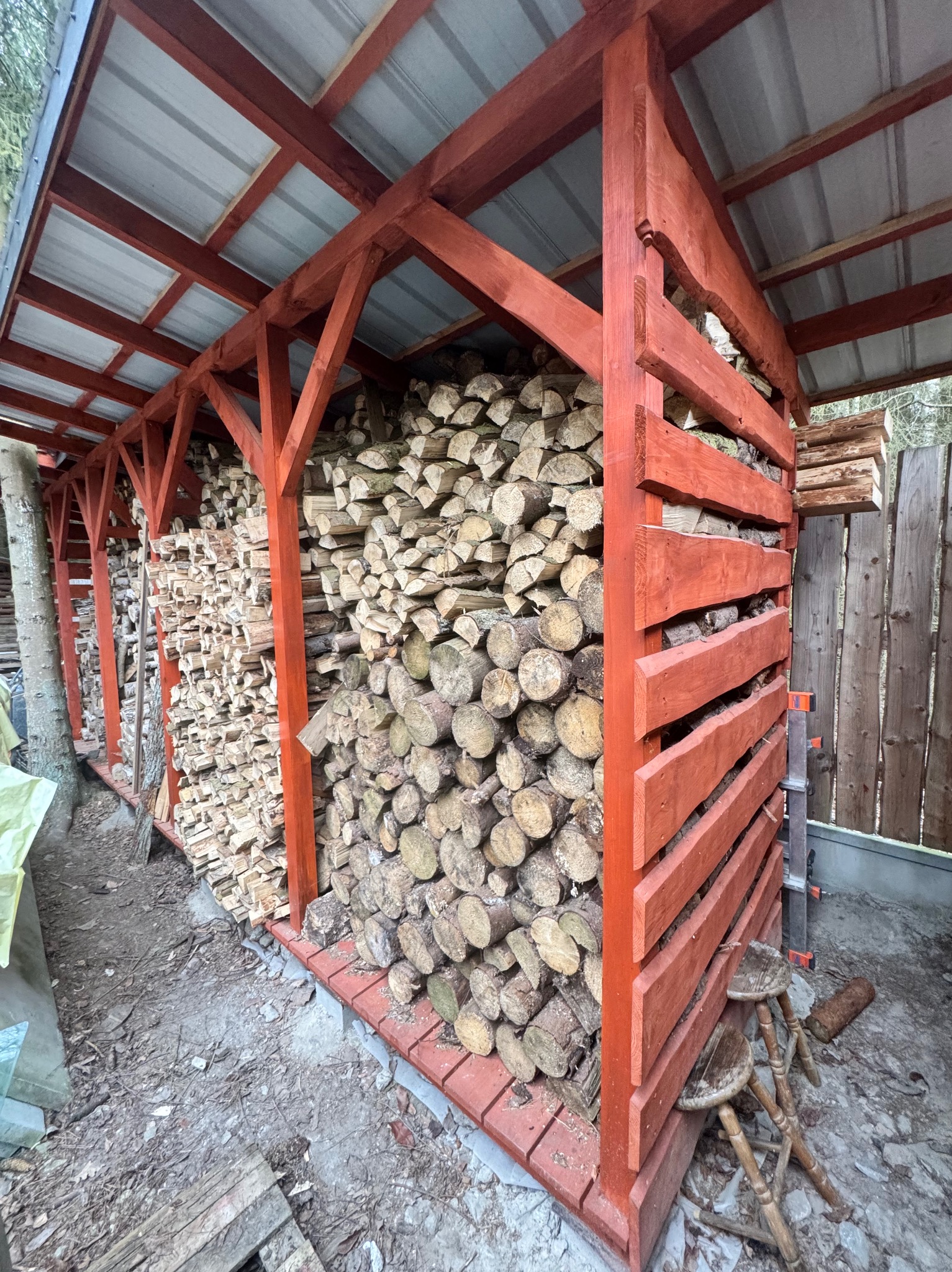 Duża drewutnia z czerwonymi, drewnianymi elementami konstrukcyjnymi i metalowym dachem, wypełniona pociętym drewnem, z dwoma stołkami obok.