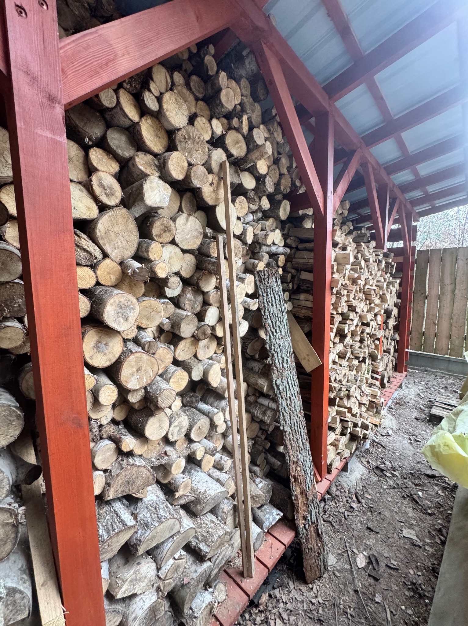 Drewniana konstrukcja z czerwonymi belkami, wypełniona ułożonymi kawałkami drewna opałowego, widok na zewnątrz konstrukcji.