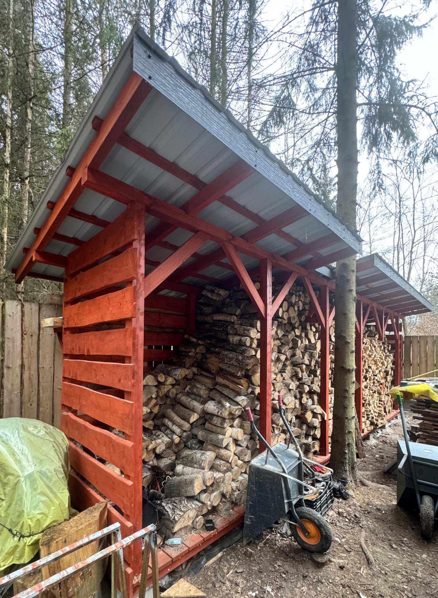 Drewniana wiata na drewno z szarą blachą falistą, wypełniona pociętymi klockami drewna. Obok taczka i narzędzia. W tle las i ogrodzenie.