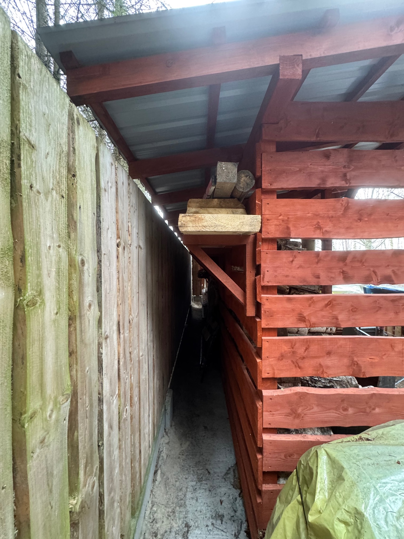 Zadaszona konstrukcja z drewnianych desek, pomalowana na czerwono, przylegająca do drewnianego płotu. Widoczny wąski przesmyk między konstrukcją a płotem.